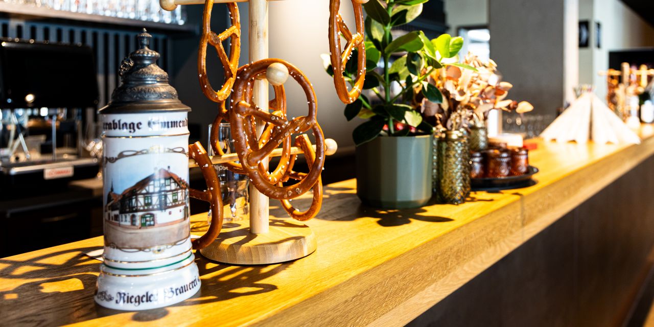 Hölzerne Theke, darauf ein traditioneller Bierkrug mit Zinndeckel und ein hölzerner Brezenständer mit aufgehängten Brezen. Daneben eine grüne Pflanze in einem Topf.